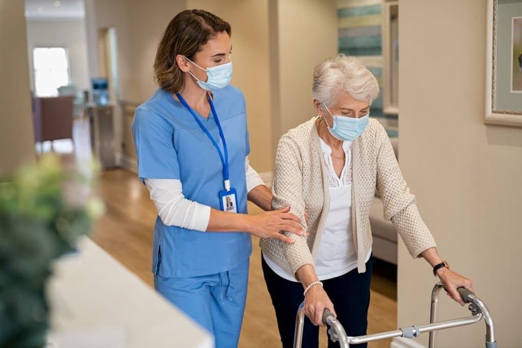 Smiling nurse with face mask helping senior woman to walk around the nursing home with walker